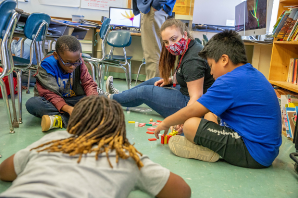 UA student sits on the floor with elementary students as they work with blocks
