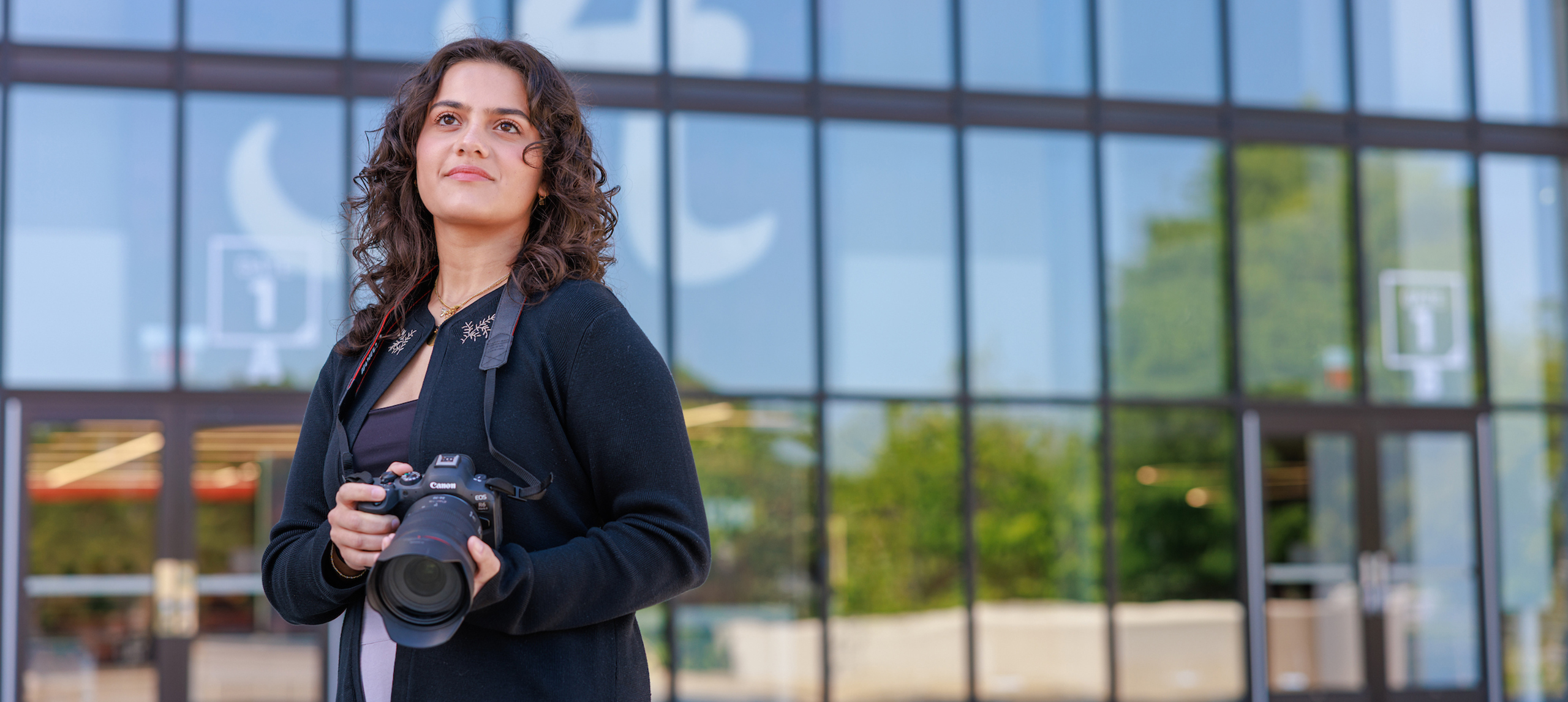 A female college students holds a camera and poses for a photo outdoors