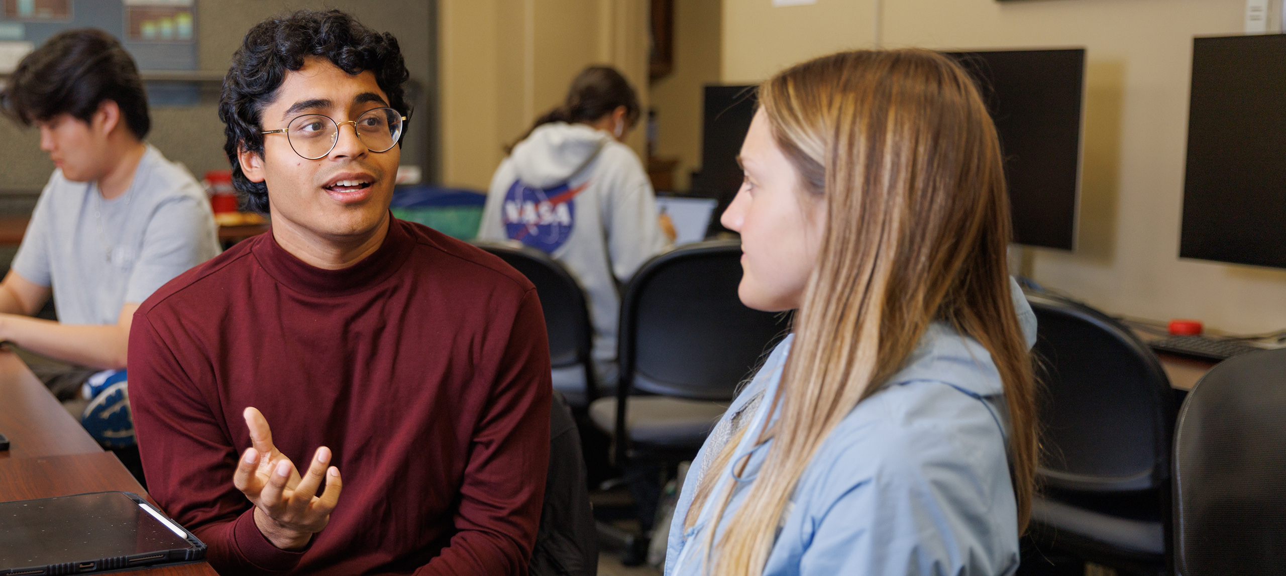 A male college student sits at a table and converses with a female classmate